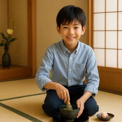 新宿・早稲田の茶道教室結空で、小学生の男の子が正座して抹茶を点てる体験風景。初心者でも安心の茶道体験で、笑顔で茶筅を使い、お隣には季節の和菓子、背景には障子と床の間の花が飾られている。