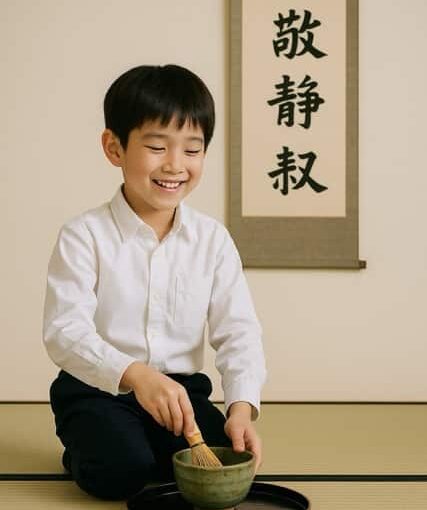 掛け軸『和敬静寂』を背景に、白いシャツを着た少年が茶筅で抹茶を点てながら笑顔を見せている写真。