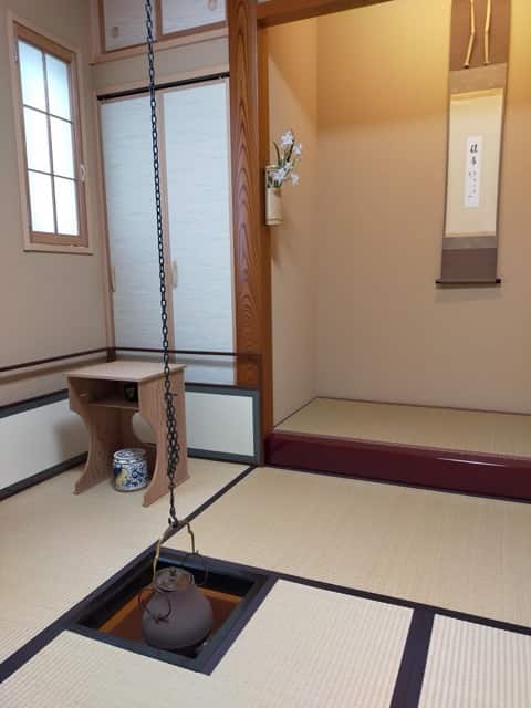 吊り釜がかけられた炉と、花が置かれた棚、床の間の掛け軸がある茶室風景。