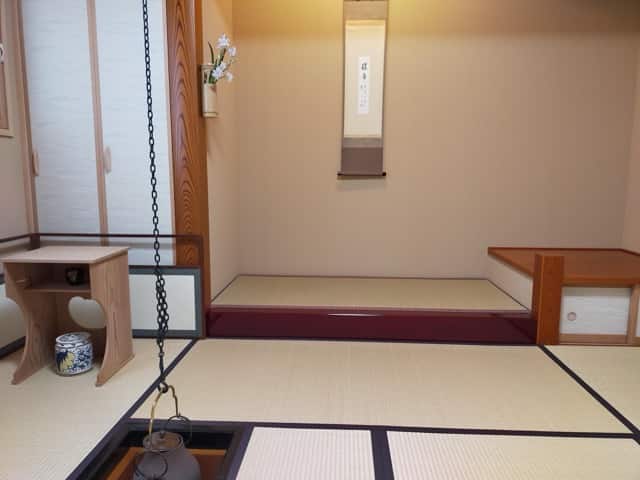 畳に切られた炉に吊り釜がかけられた茶室。床の間には花と掛け軸が飾られている。