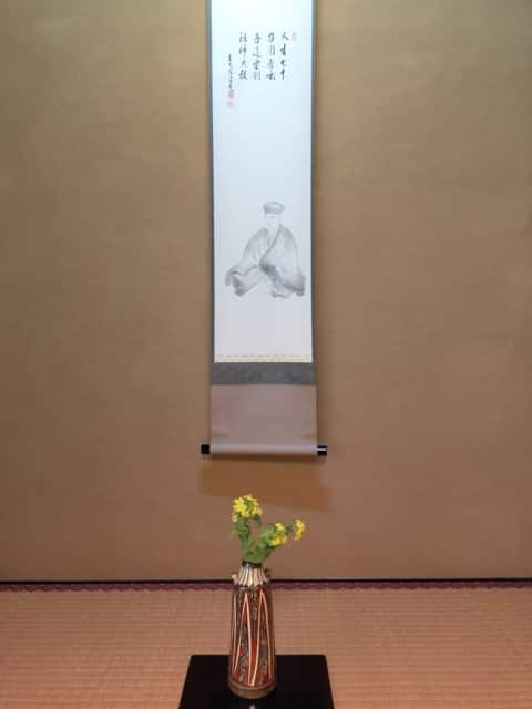 利休居士の掛物の前に、琉球花入に生けた菜の花を飾った床の間の写真。春の利休忌を静かに表す一枚。
